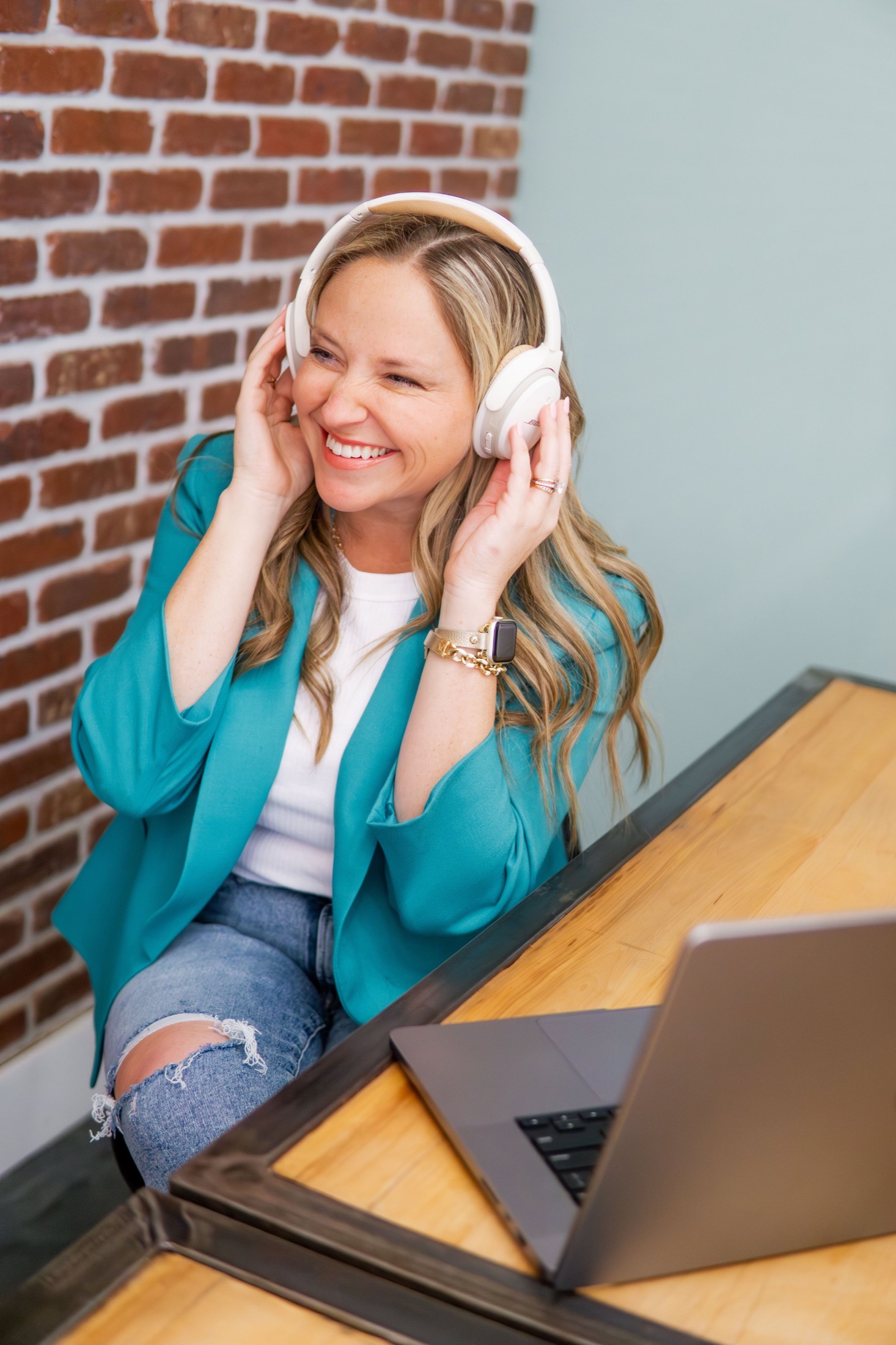 A smiling woman wearing a teal blazer and ripped jeans, sitting at a wooden desk with a laptop, wearing headphones, near a brick wall.